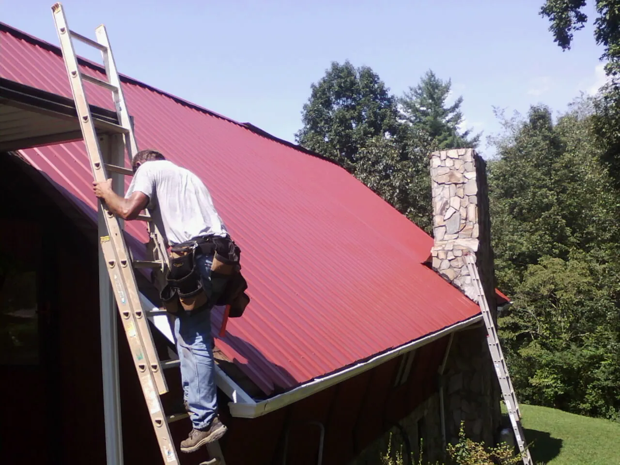 Roofer on a ladder beside a red metal roof and stone chimney — Soffit & Fascia Repair by Roofing Brothers in Ashton